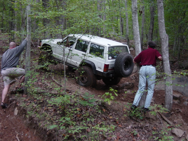 White Jeep Cherokee XJ climbing a steep, wooded trail with two people nearby