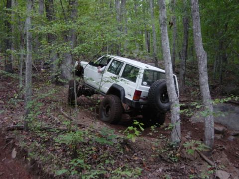 1991 Ford Bronco - A white Jeep Cherokee navigates a challenging off-road trail through a dense forest.