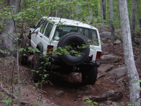 1991 Ford Bronco - A white Jeep Cherokee XJ navigates a challenging rocky trail in a wooded area.