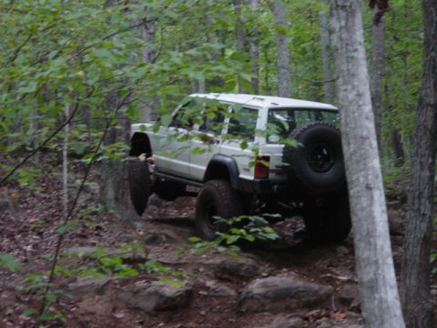 1991 Ford Bronco - A white Jeep Cherokee XJ navigates a rocky, wooded trail with its spare tire mounted on the back.