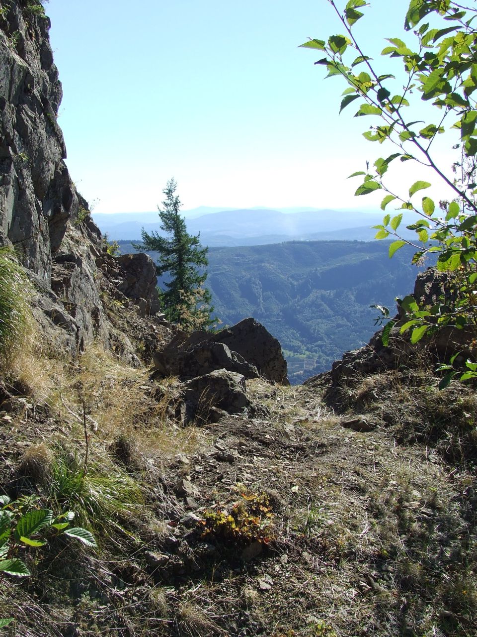 Rocky mountain path overlooking a valley with distant hills under a clear blue sky