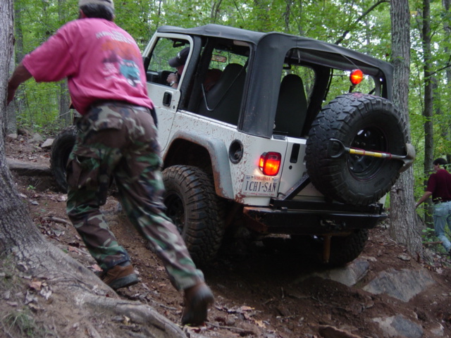 White Jeep Wrangler climbing a rocky trail with a person pushing from behind. Spare tire mounted on the back
