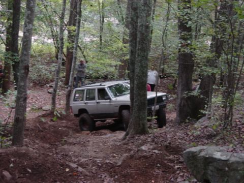 1991 Ford Bronco - A Jeep navigates a challenging off-road course through a dense forest with onlookers present.
