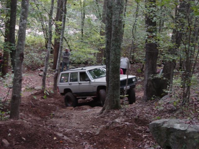 Silver Jeep Cherokee XJ climbing a dirt trail in a wooded area. Two people stand nearby