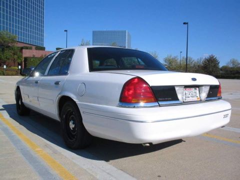 1998 Ford Crown Victoria - A white sedan is parked in a lot with a modern building in the background. The rear of the car is visible.