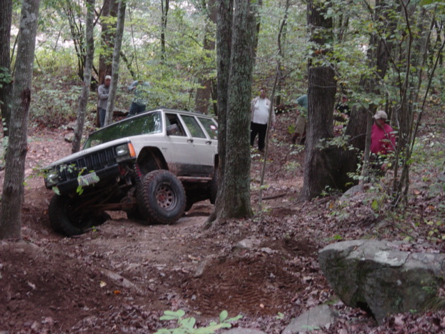 White Jeep Cherokee XJ off-roading on a dirt trail, angled left, with large tires and lifted suspension