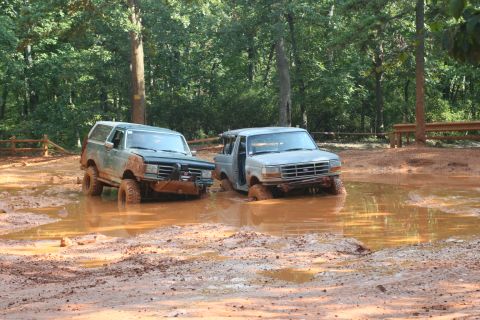 1990 Ford Bronco - Two Ford Broncos are stuck in a muddy off-road area, surrounded by trees.
