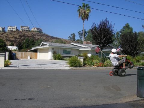 1900 Honda Big Red - A person performs a wheelie on a three-wheeled vehicle in a suburban street, with a house and palm tree in the background.