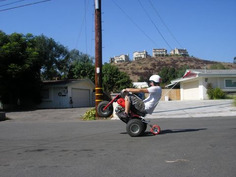1900 Honda Big Red - Man performs a wheelie on a red three-wheeled ATV on a sunny day in a suburban neighborhood.