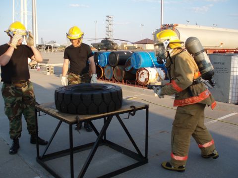 1981 Ford Bronco - Firefighters prepare for a training exercise involving a tire and barrels, with aircraft and a helicopter in the background.