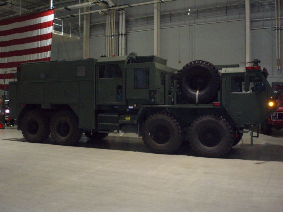 Green military truck with multiple axles and a spare tire mounted on the rear