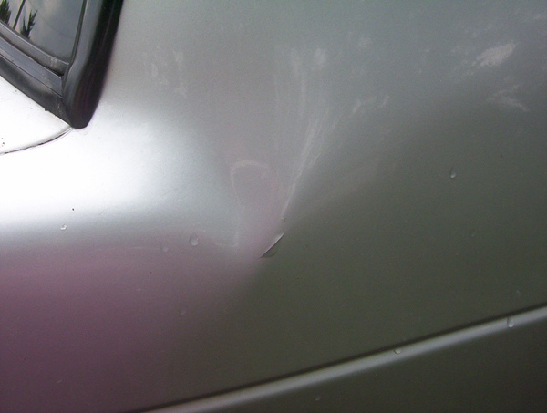Close-up of a dent and paint chip on a silver car body panel with water droplets