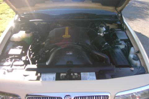 1996 Mercury Grand Marquis - Overhead view of a V8 engine bay in a light tan/cream sedan, showing a black plastic engine cover marked "V8" with a yellow decal, coolant…