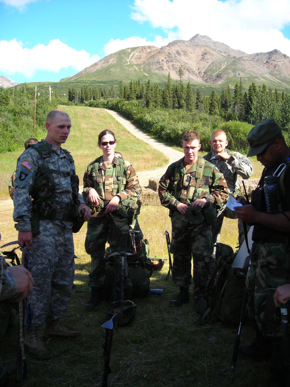 Group of soldiers in camouflage uniforms standing in a grassy field with mountains and trees in the background