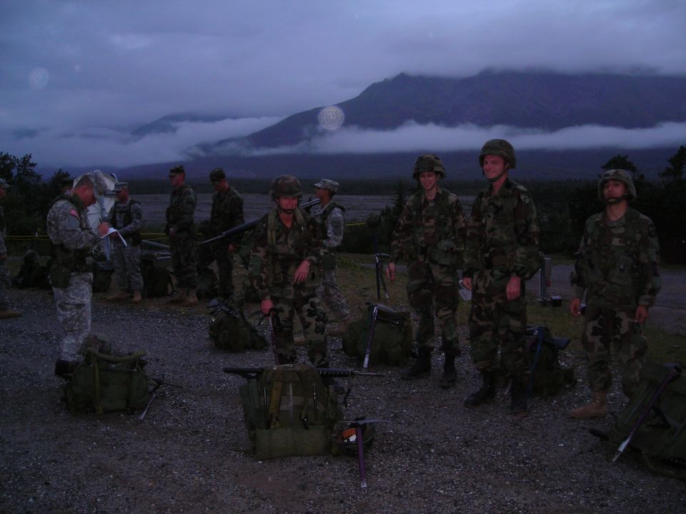 Group of soldiers in camouflage uniforms and helmets stand around with their gear, including backpacks and rifles, in front of a cloudy mountain range