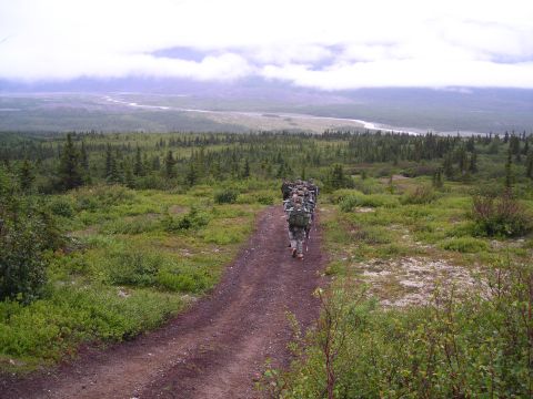 2003 Jeep TJ - A group of soldiers trek along a dirt path in a vast, natural landscape with trees and a distant river under a cloudy sky.