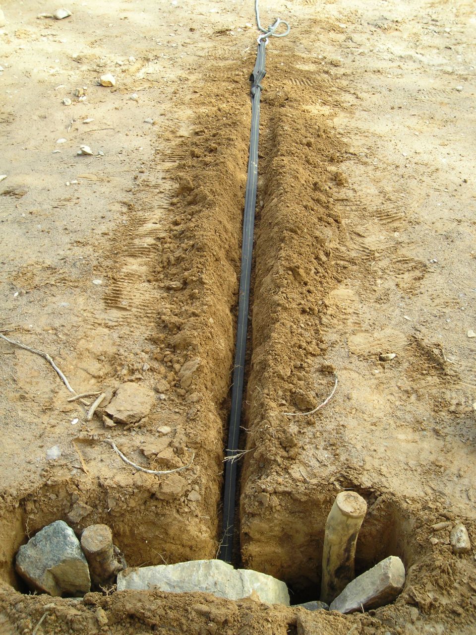 Trench dug in dirt with a cable running down the middle. Wooden stakes and rocks are visible at the bottom