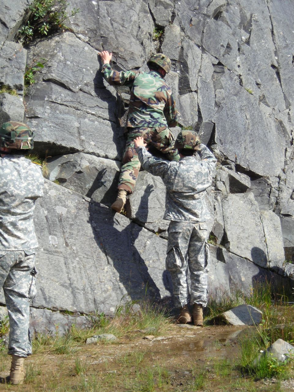 Soldiers in camouflage uniforms practice rock climbing on a steep, rocky incline