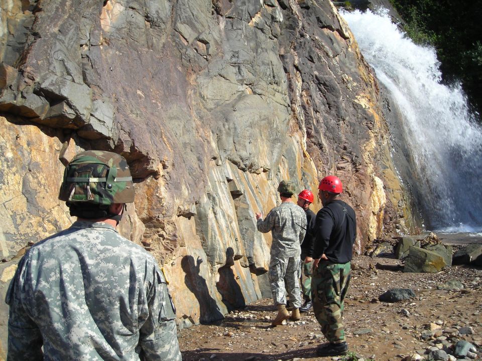 Soldiers and a civilian in hard hats look at a rock formation next to a waterfall