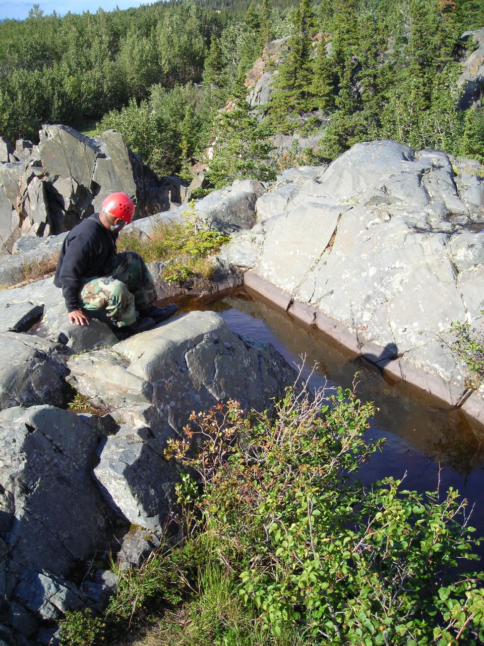 Man wearing a red helmet and camouflage pants kneels by a narrow rock pool with trees and rocky terrain in the background