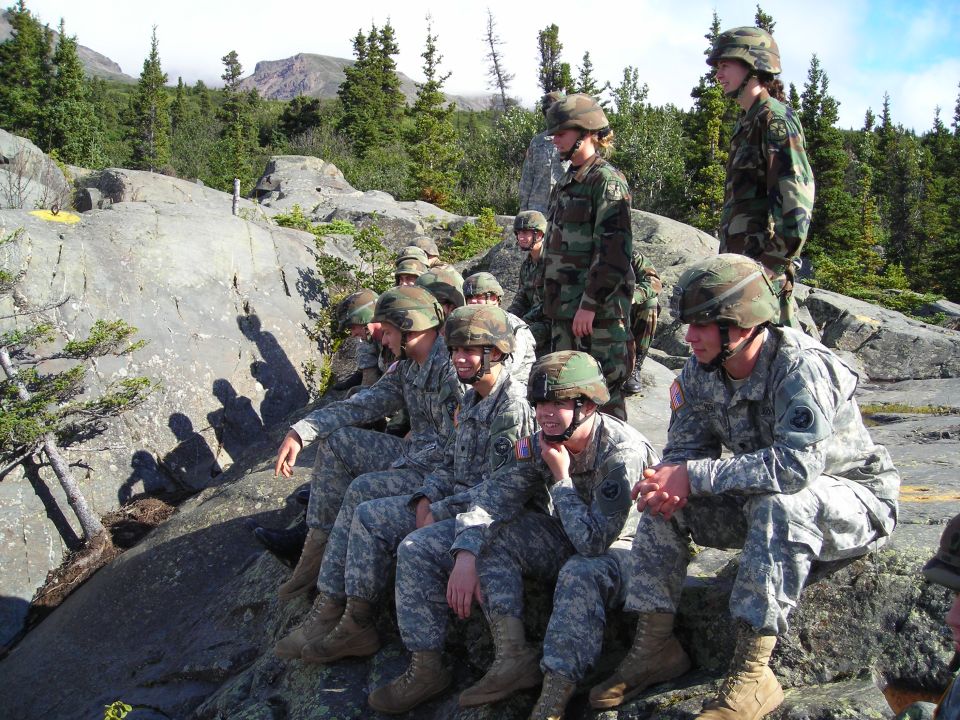 Group of soldiers in camouflage uniforms and helmets sitting on rocks outdoors, with trees and mountains in the background