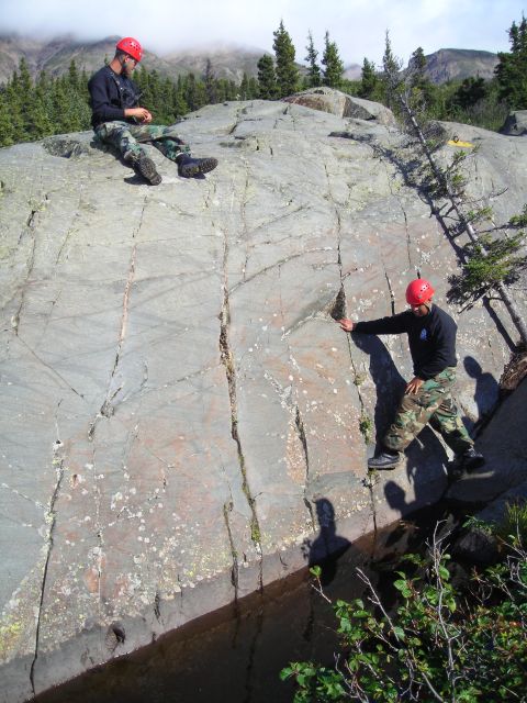2003 Jeep TJ - Two individuals in climbing gear are positioned on a large rock face, with trees and mountains visible in the background.
