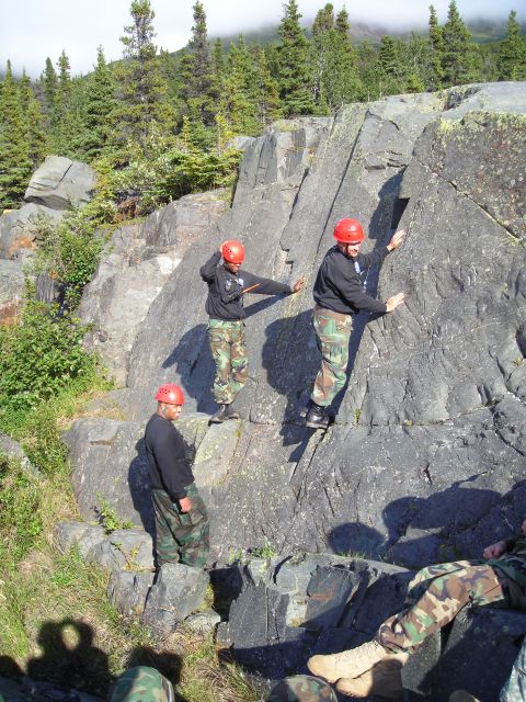 2003 Jeep TJ - People in helmets and camouflage gear ascend a steep rock formation in a wooded area.
