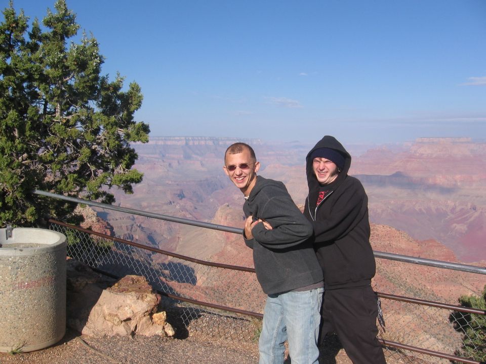 Two guys posing for a photo at the Grand Canyon overlook. A tree is on the left, and a concrete structure is in the foreground