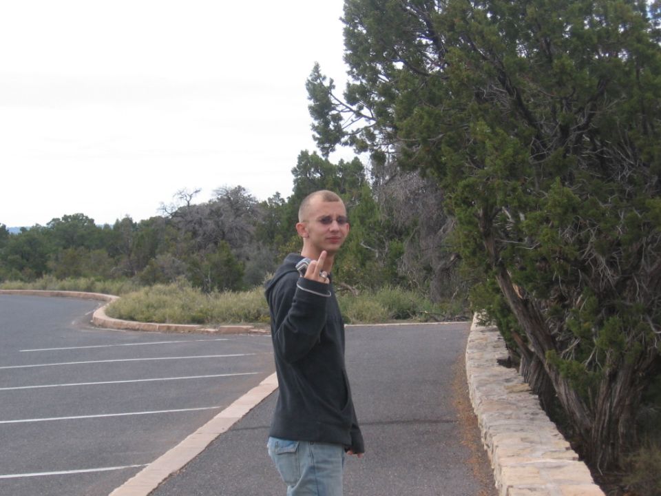 Man with shaved head and beard, wearing a dark hoodie and jeans, gives the middle finger while standing on a paved road next to trees