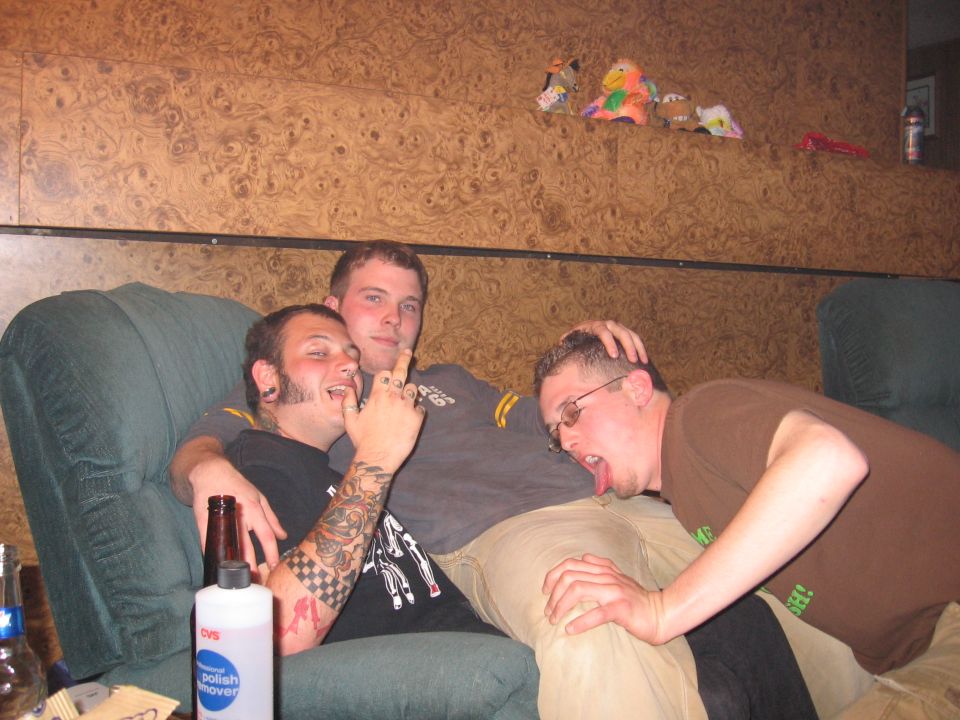 Three men on a green couch, one with tattoos and piercings, another with glasses licking the leg of the third man