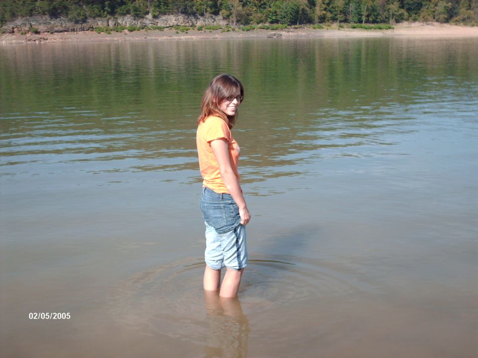 Woman in orange shirt and denim shorts stands in shallow water near a tree-lined shore