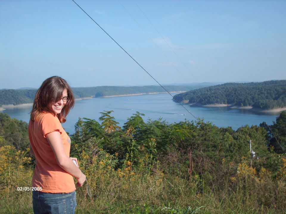 Woman in orange shirt and jeans stands on a hill overlooking a wide lake with tree-covered shores under a clear blue sky