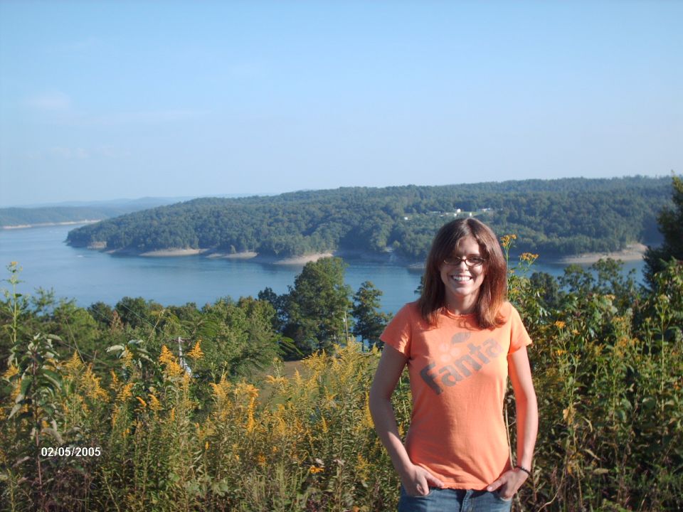 Woman in orange t-shirt stands in front of a lake and wooded hills