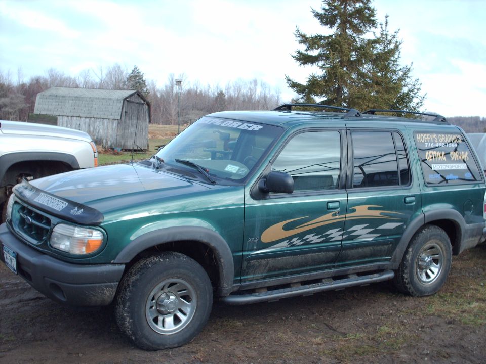 Green SUV with racing decals and roof rack, parked on dirt. A silver truck is partially visible to the left