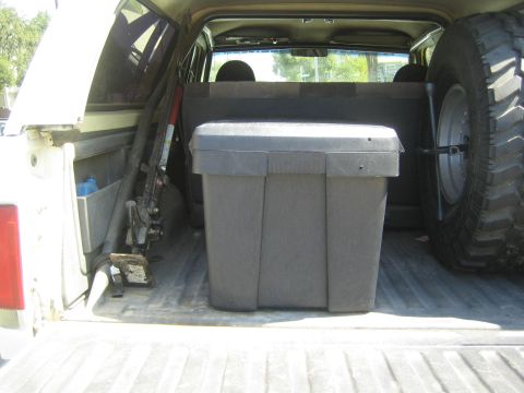 1990 Ford Bronco - gray storage tote and a floor jack sit in a pickup bed alongside a large spare off-road tire.