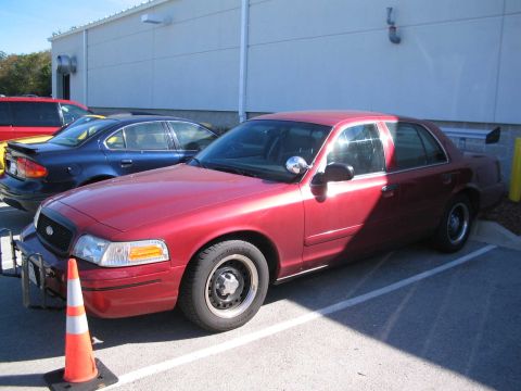 2001 Ford Crown Victoria - A red 2001 Ford Crown Victoria sedan is parked next to other vehicles, with a traffic cone in the foreground.