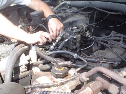 1984 Ford Bronco - Hands work on the carburetor in a cluttered engine bay.