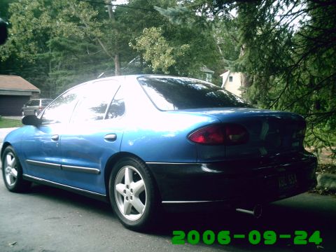 1996 Chevrolet Cavalier - The rear of a blue sedan is visible, parked in a residential area with trees and houses in the background.