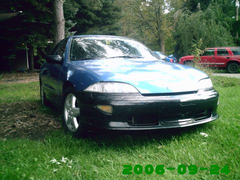 1996 Chevrolet Cavalier - A blue car with a contrasting black bumper is parked on a grassy area, with a red SUV in the background.
