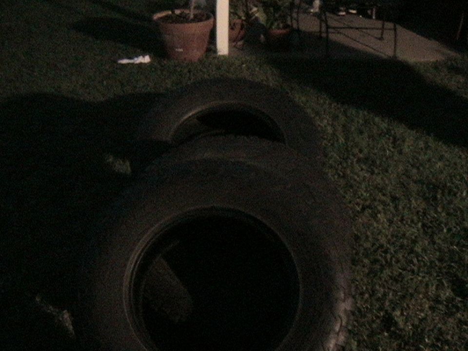 Two black tires lying on green grass, one partially obscuring the other. A potted plant and chair are visible in the background