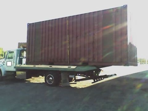 1992 Ford Bronco - Flatbed truck transports a large shipping container under a bright sky.