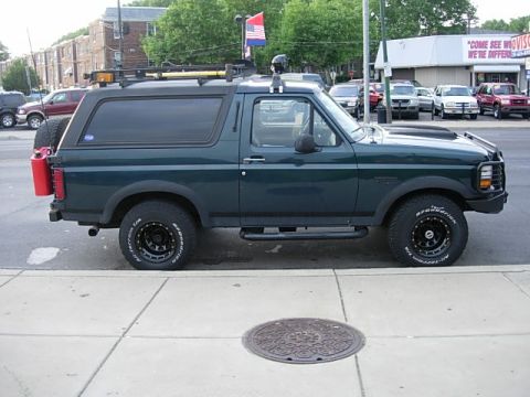 1993 Ford Bronco - A dark green Ford Bronco is parked on the side of the road with a roof rack and spare tire mounted on the rear.