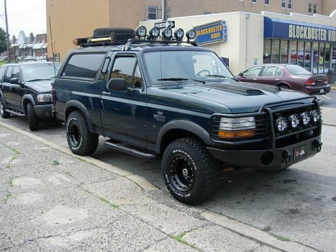 1993 Ford Bronco - A modified Ford Bronco and an SUV are parked on a street in front of a Blockbuster Video store.