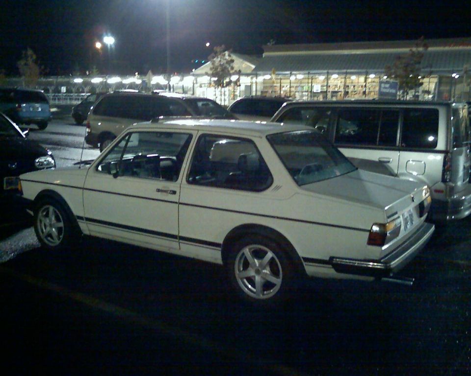 White Volkswagen sedan parked at night, showing its side and rear quarter panel. The car has a black stripe along its side and aftermarket wheels