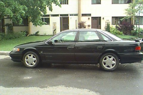 1993 Ford Taurus - A black 1993 Ford Taurus sedan is parked on a wet road in front of a building.