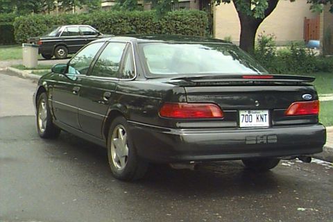 1993 Ford Taurus - A black sedan is parked on a wet street, with another car visible in the background.