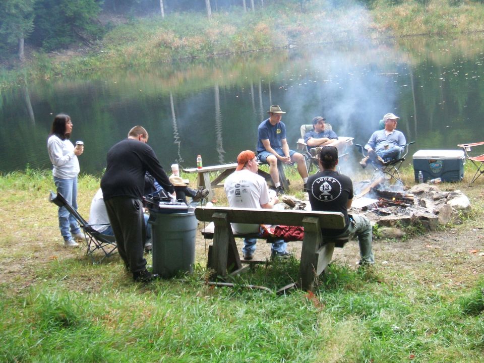 Campfire scene by lake with people sitting on benches and chairs