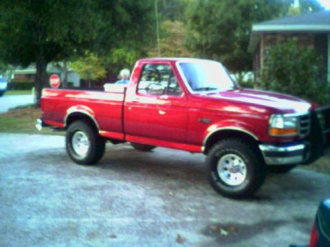 1992 Ford F-150 - Red Ford F-Series regular-cab short-bed pickup parked at an angle on a gravel driveway, shown from the passenger side with the front wheels…