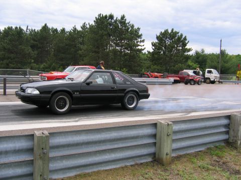 1989 Ford Mustang - A black 1989 Ford Mustang hatchback performs a burnout on a drag strip, with other vehicles visible in the background.