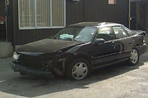 1993 Ford Taurus - This black 1993 Ford Taurus sedan shows significant damage to its front end, including the bumper and hood.